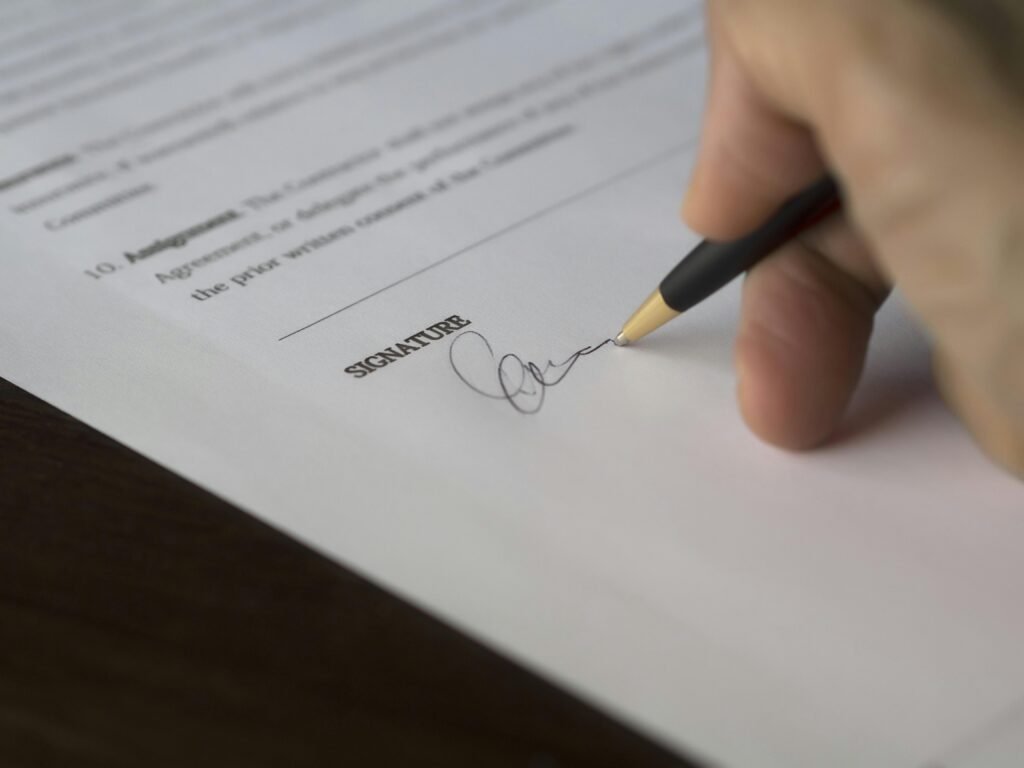 A hand signs a formal contract with a pen on a wooden desk.