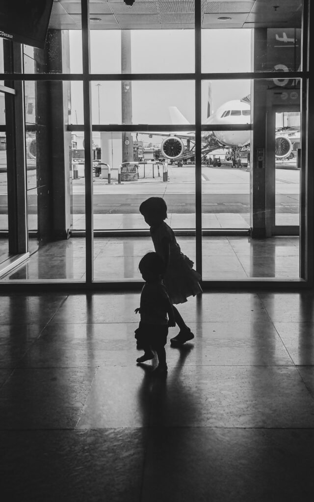 Two children silhouette walking in an airport terminal with an airplane in the background.