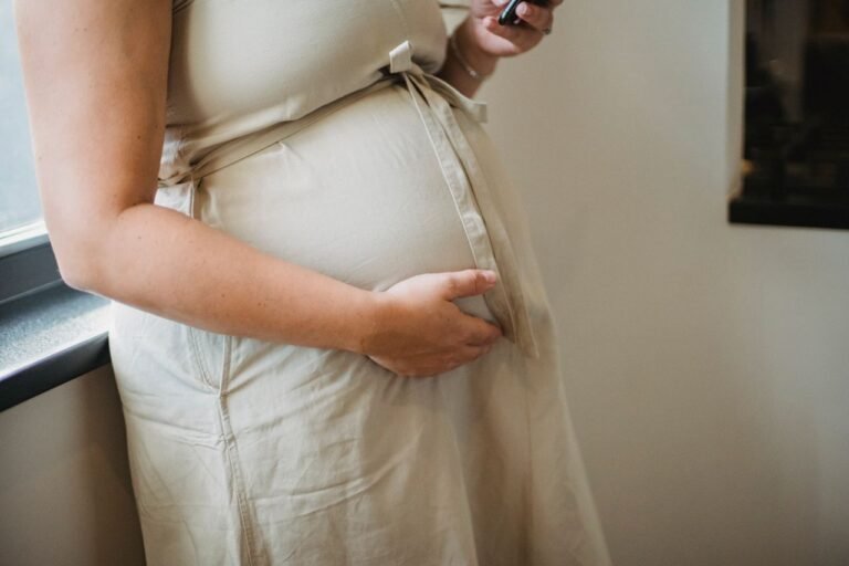 Crop unrecognizable pregnant female standing near window and checking notification on cellphone in room